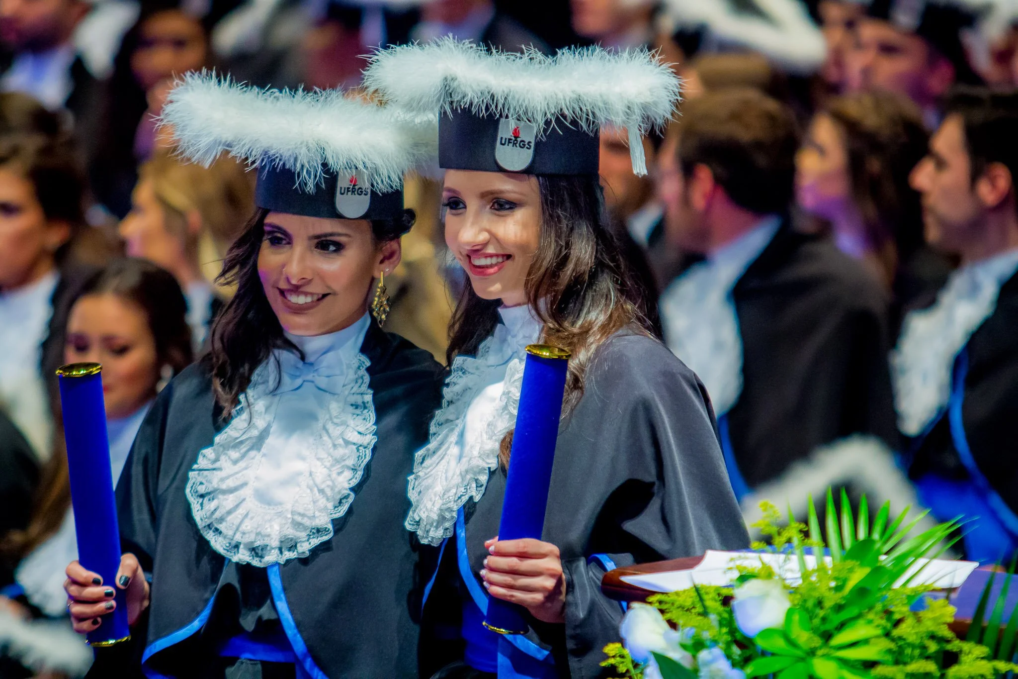 Duas mulheres em vestimentas acadêmicas comemorando a formatura, usando chapéis com plumas brancas, segurando diploma em mãos, em uma cerimônia de formatura universitária.