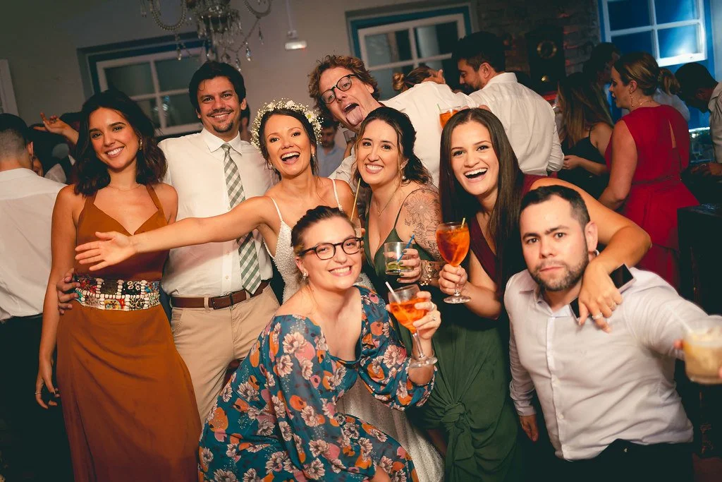 Grupo de pessoas celebrando, sorrindo, segurando drinks coloridos, em uma festa com decoração elegante.