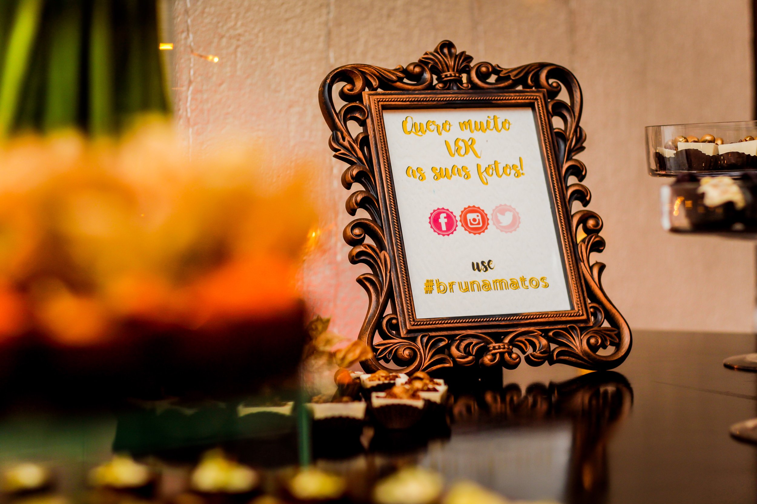 Painel decorativo com mensagem escrita em português,pedra ornamentada ao redor, em um evento social, com doces ao redor e vasos de flores na frente.