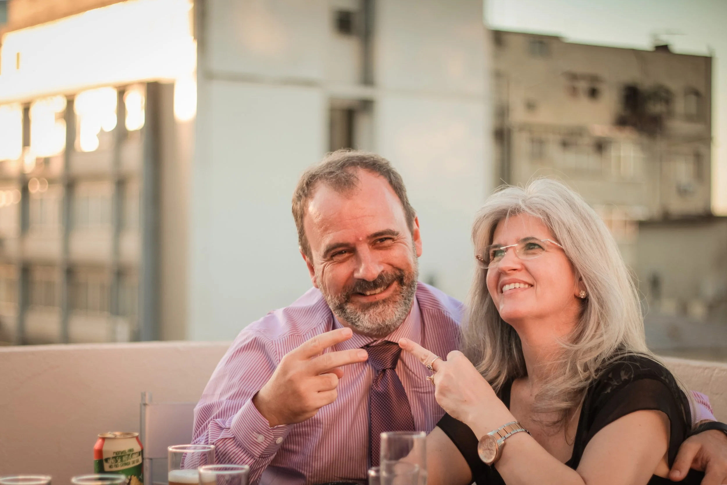 Casal sorridente celebrando juntos em um evento ao ar livre, com bebidas na mesa ao fundo.