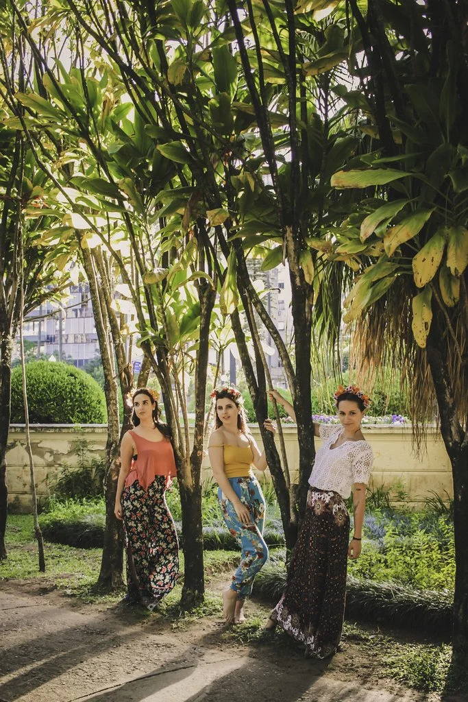 Três mulheres com roupas florais e coroas de flores posando em um jardim sob árvores.