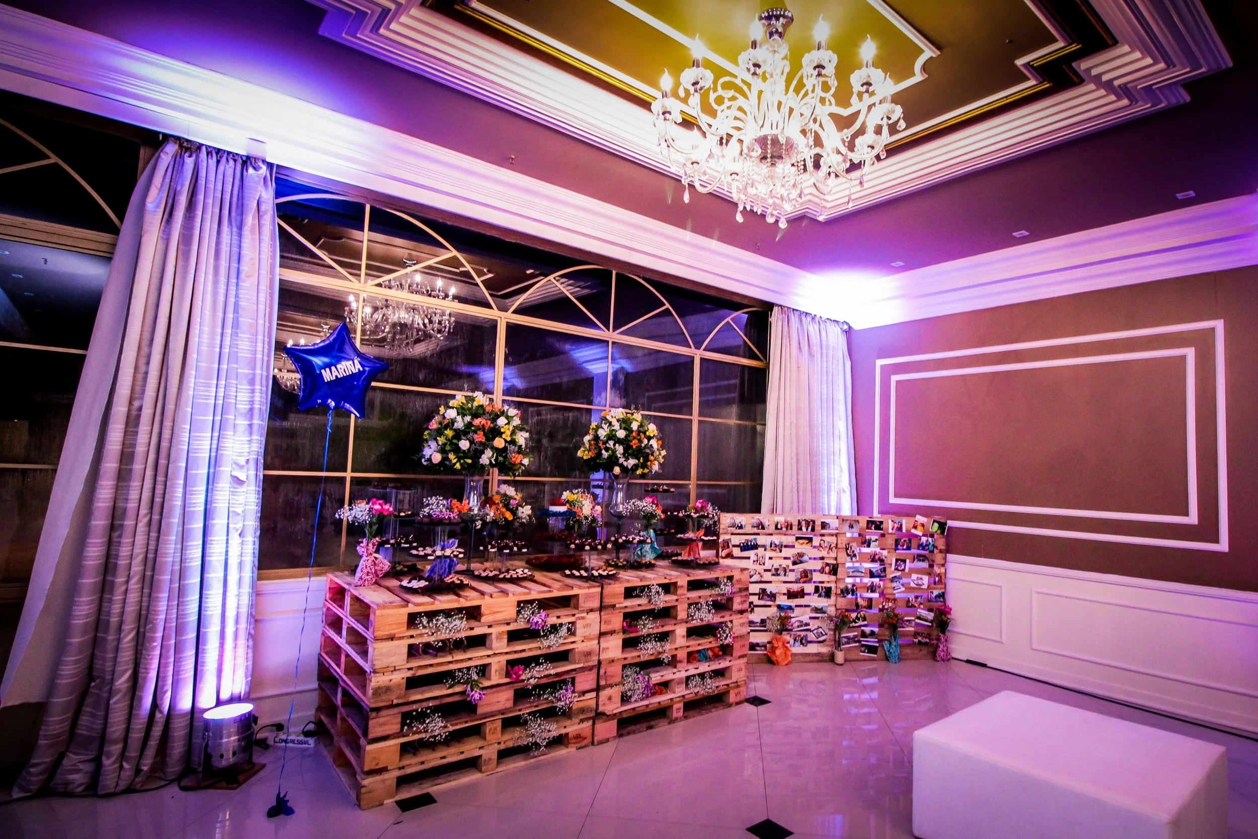 Sala decorada com flores, quadros e uma luminária cristal, com cortinas e janela grande ao fundo, iluminação suave com luzes coloridas, espaço para comemoração.