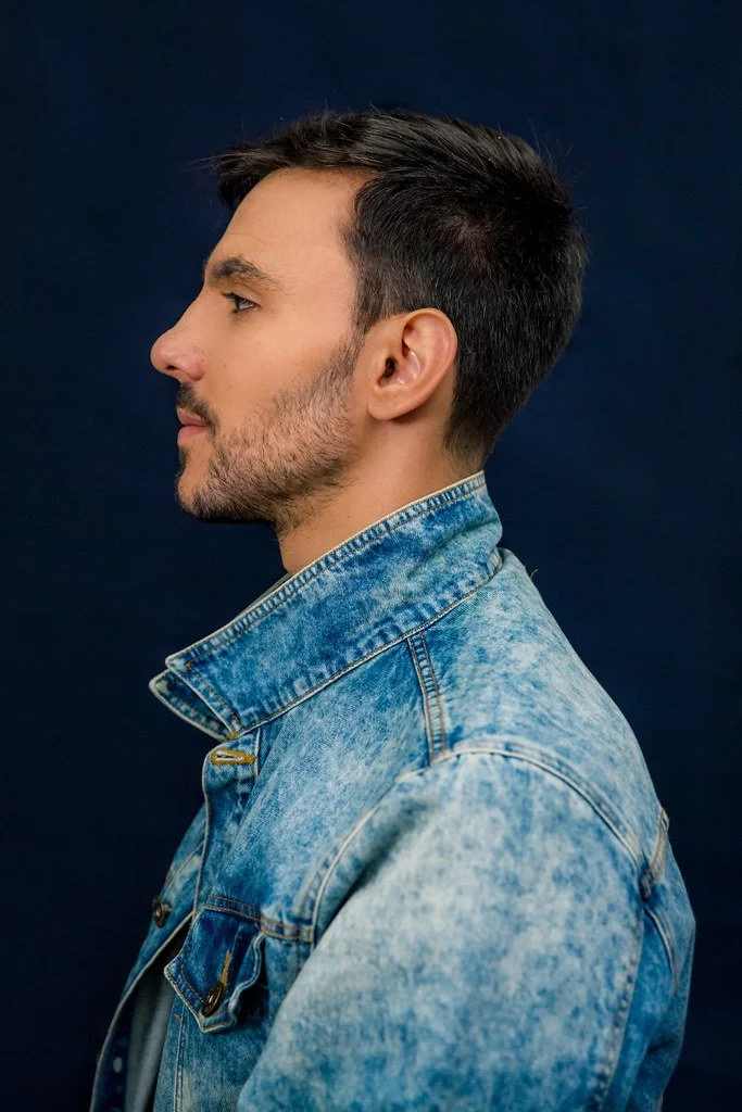 Retrato de perfil de um homem de cabelo castanho escuro, barba por fazer, usando jaqueta jeans azul claro contra fundo escuro.