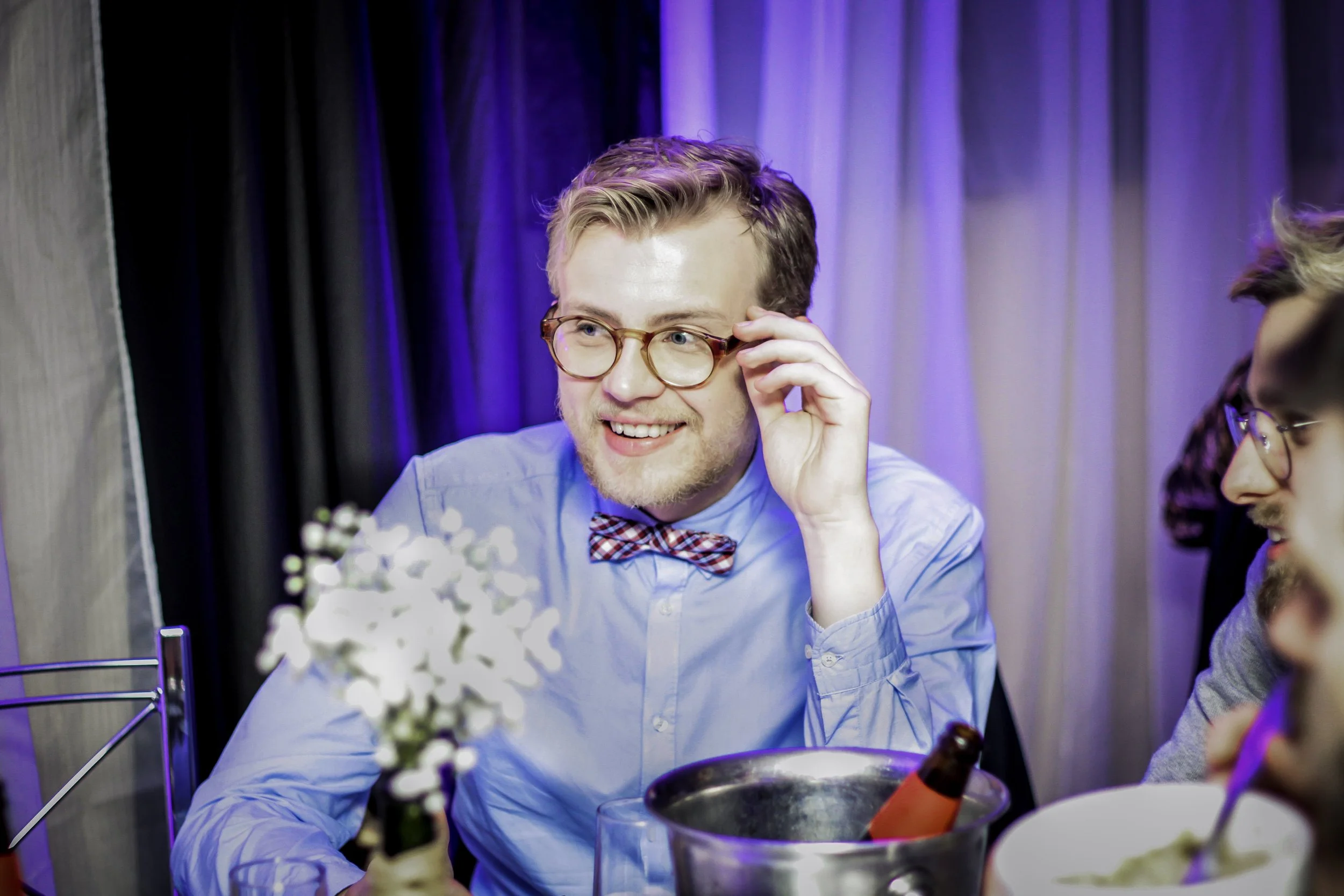 Homem sorridente com óculos redondos, camiseta azul claro, gravata borboleta xadrez, em festa, sentados à mesa com flores, bebidas e comida, fundo com cortinas.