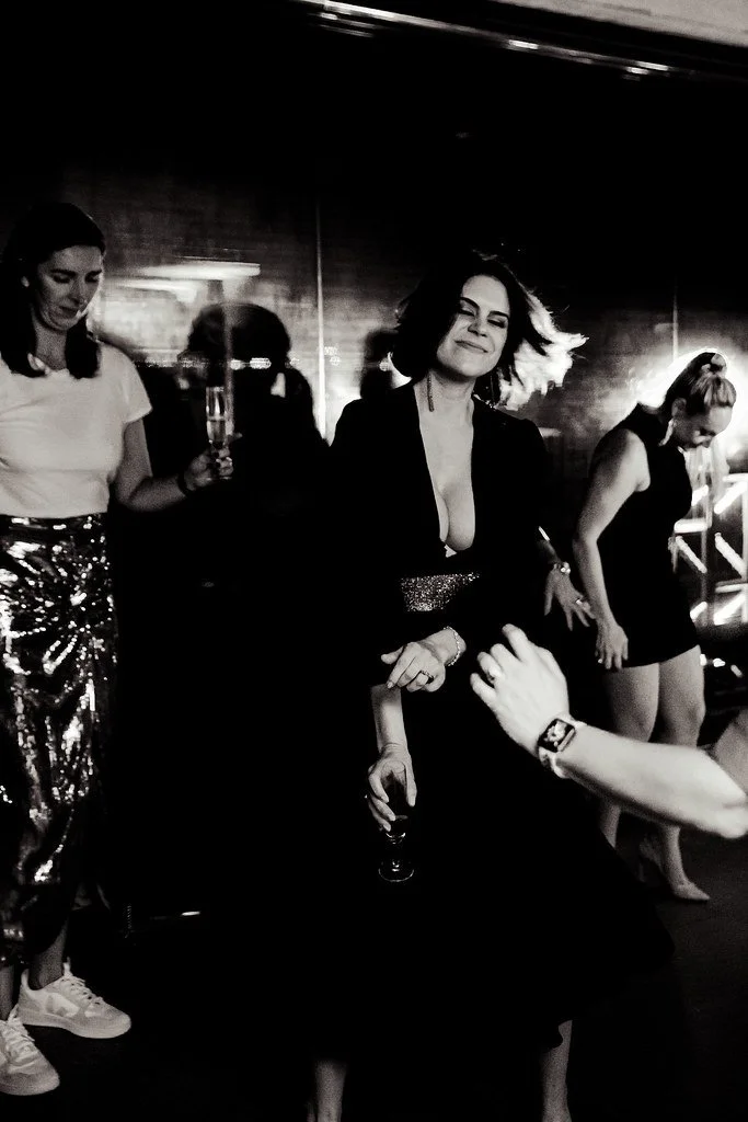 Mulheres dançando e curtindo em uma festa ou balada, ambiente escuro com luzes de fundo, uma mulher ao centro com cabelo ao vento, vestindo vestido preto.