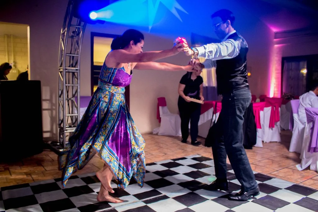 Casal dançando no chão de xadrez em uma festa, com mesas decoradas ao fundo, sob iluminação colorida.