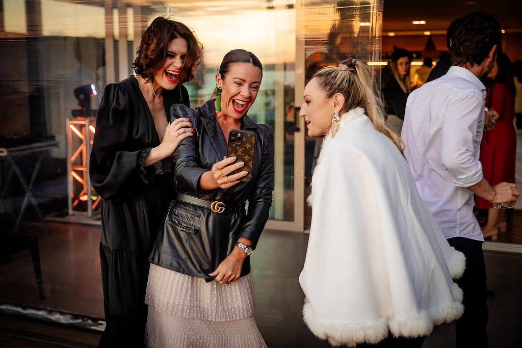 Três mulheres sorrindo enquanto olham um celular numa festa.