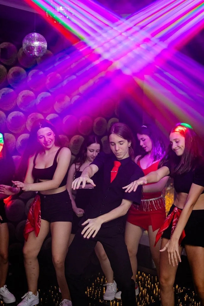 Grupo de pessoas dançando em uma festa sob luzes coloridas e um espelho espiral no fundo.