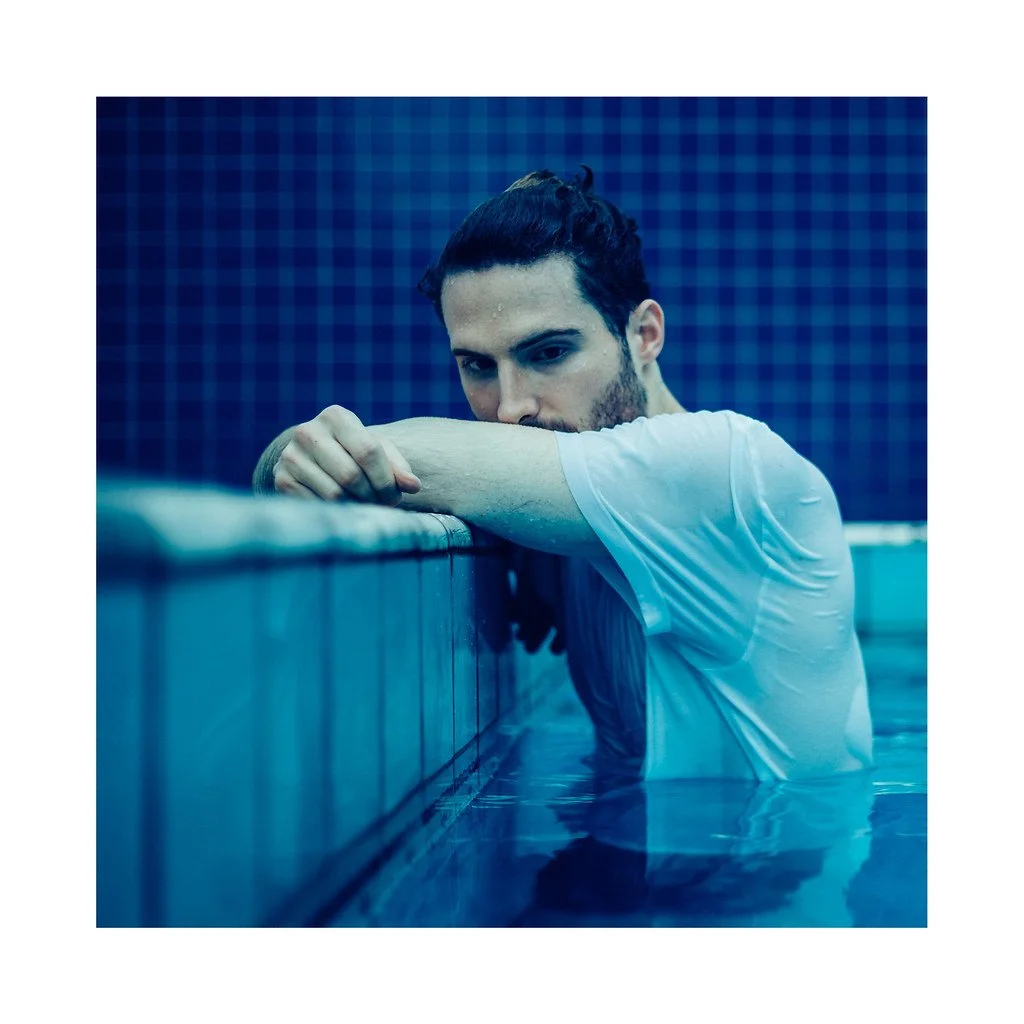 Homem com cabelo escuro preso, de barba, usando camiseta branca, apoiado na borda de uma piscina, com fundo de azulejos azuis, olhando para a câmera.