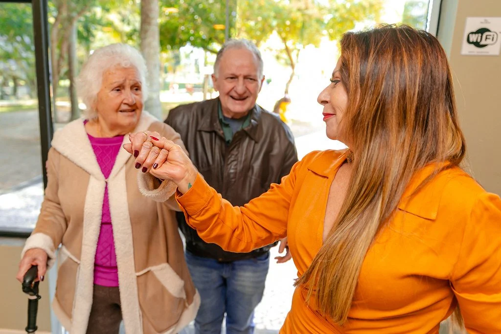 Mulher idosa recebendo a mão de uma jovem com um sorriso, enquanto um homem e uma mulher idosa observam sorrindo ao fundo.