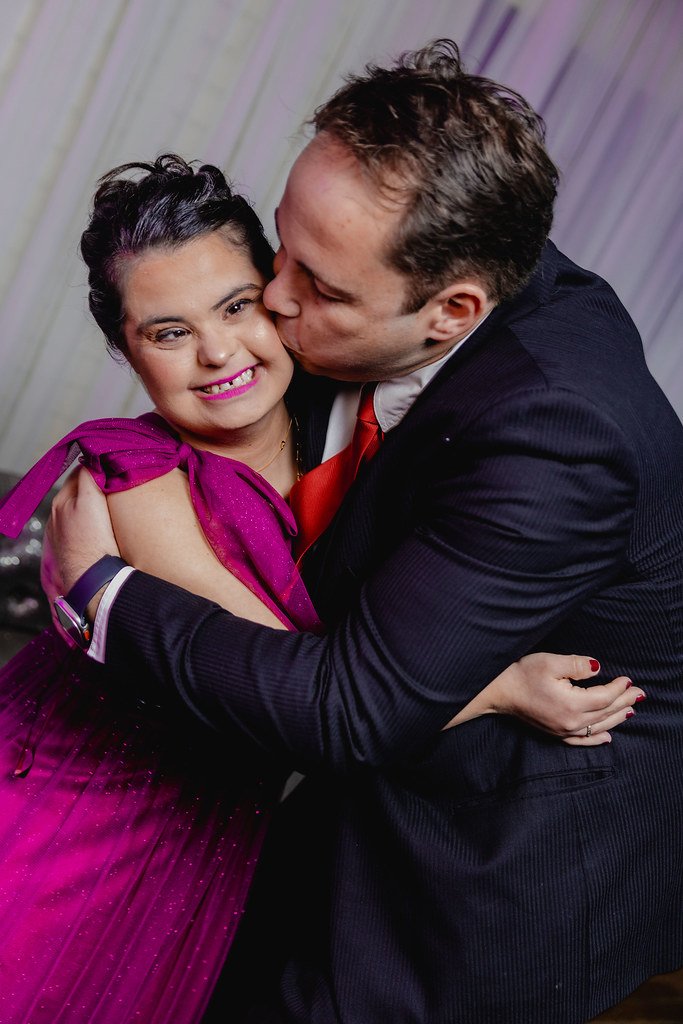 Homem e mulher se abraçando carinhosamente em uma festa, ela veste um vestido rosa com detalhes brilhantes e maquiagem vibrante, ele usa terno preto com camisa branca e gravata vermelha.