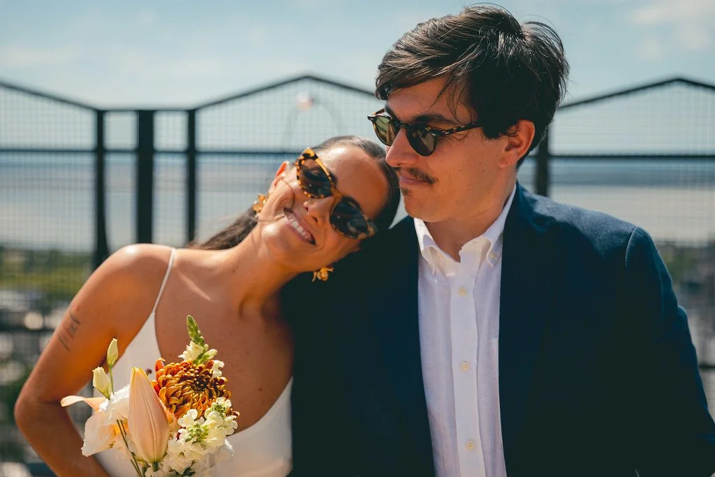 Casal sorridente em um momento íntimo ao ar livre, com a mulher segurando um buquê de flores e usando óculos grandes, enquanto o homem usa óculos escuros e veste terno escuro, com uma grade de fundo.
