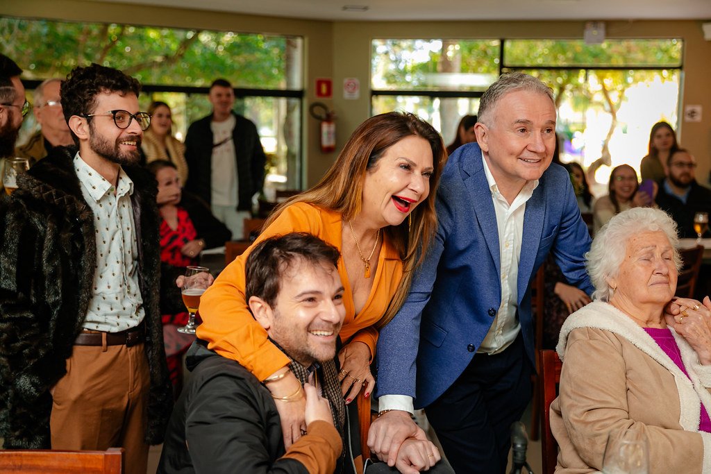 Grupo de pessoas sorrindo em um evento social, incluindo uma mulher de cabelo castanho e cabelo liso com blusa laranja, um homem de cabelo grisalho com terno azul, uma mulher idosa de cabelo branco com blusa rosa, e outras pessoas ao fundo em um ambi