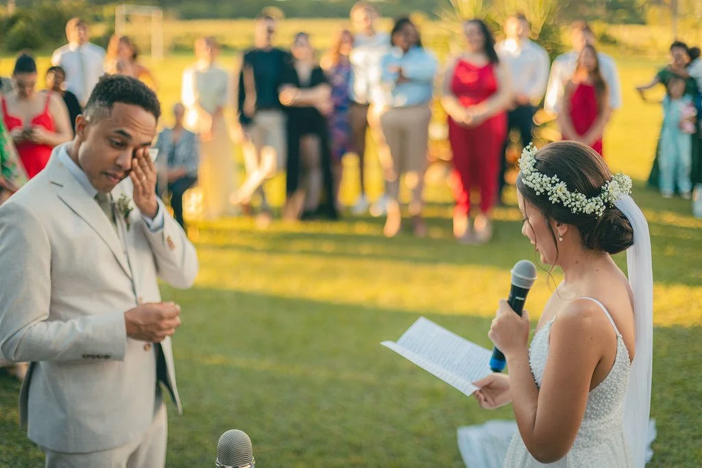 Noivos trocando votos ao ar livre, com convidados assistindo ao fundo.