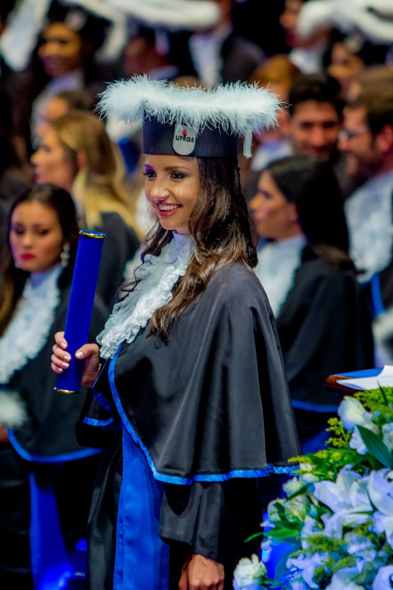 Mulher sorridente em traje de formatura, usando capelo com detalhes de penas brancas, segurando diploma azul, em cerimônia com várias pessoas ao fundo.