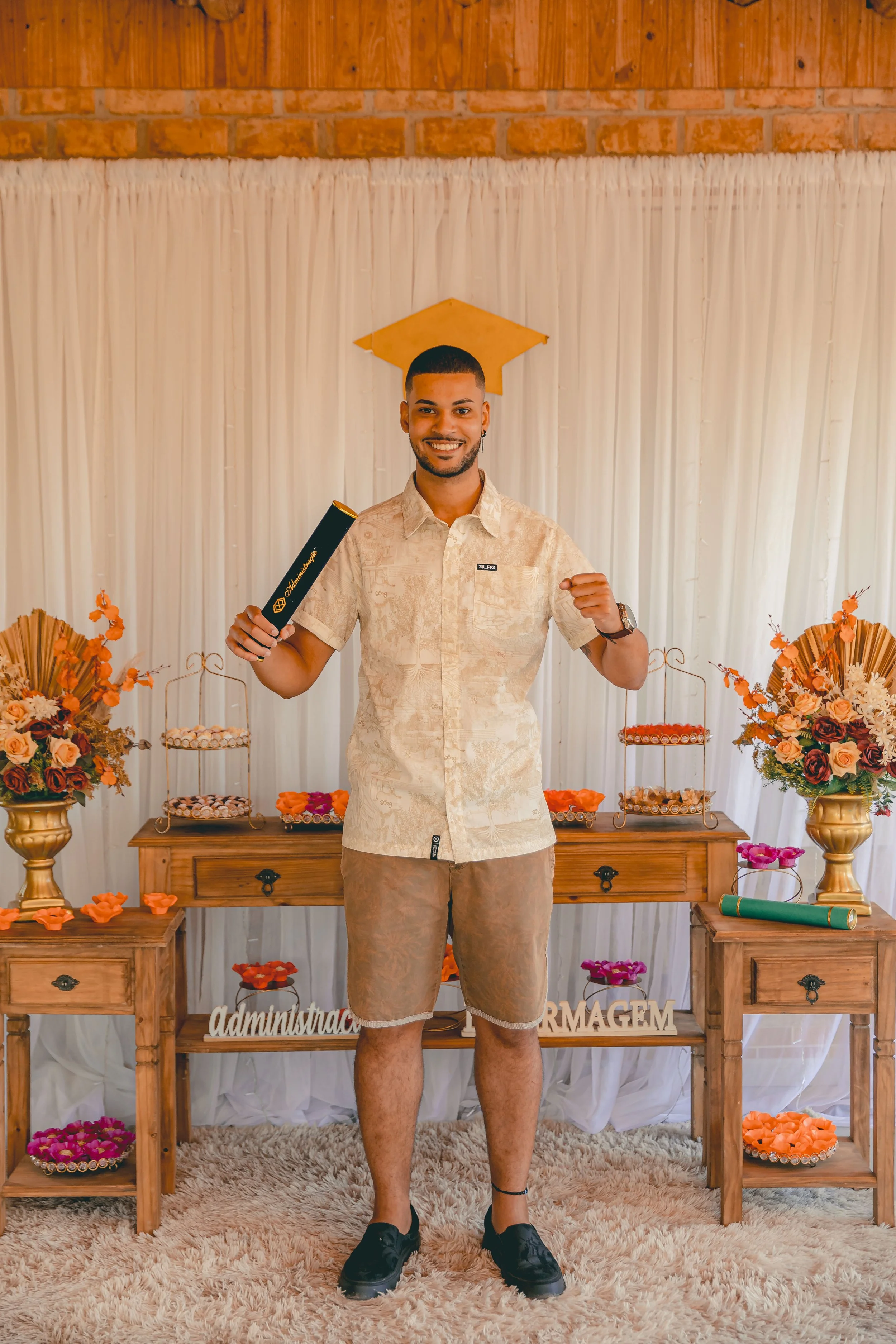 Homem sorridente de pé em frente a uma decoração de festa de formatura, segurando um diploma, com um chapéu de formatura na cabeça e uma mesa decorada ao fundo com flores, doces e letras decorativas.