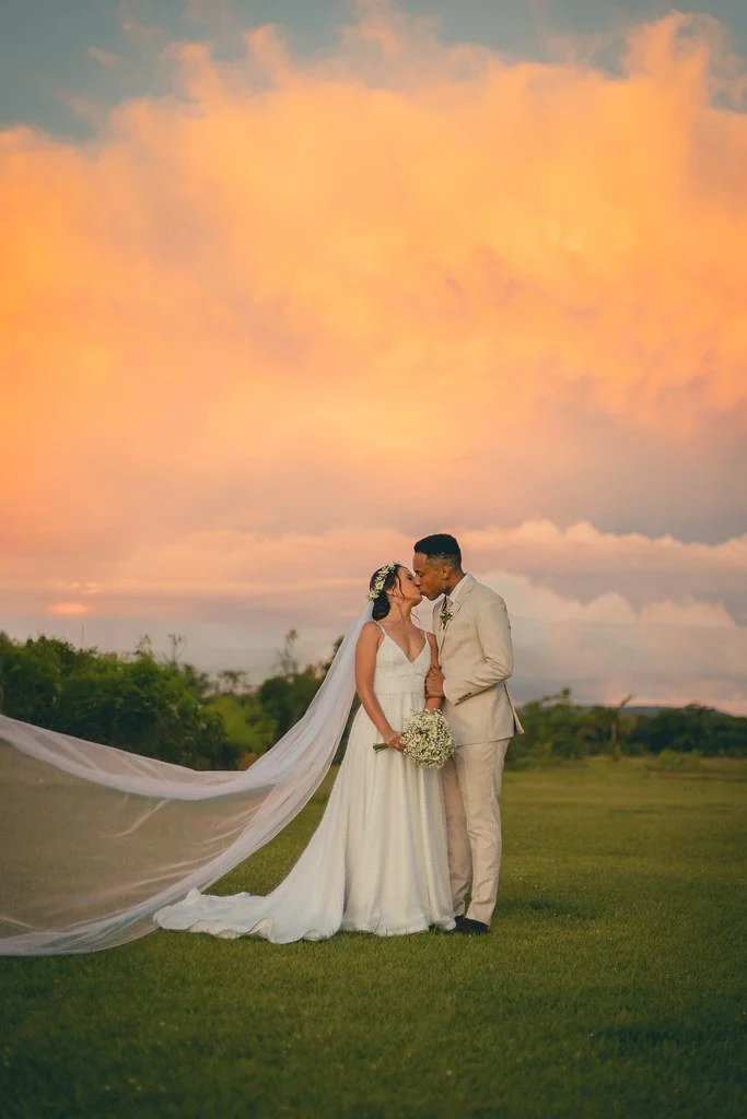 Casal de noivos se beijando ao pôr do sol, ao ar livre, com céu colorido de nuvens alaranjadas.