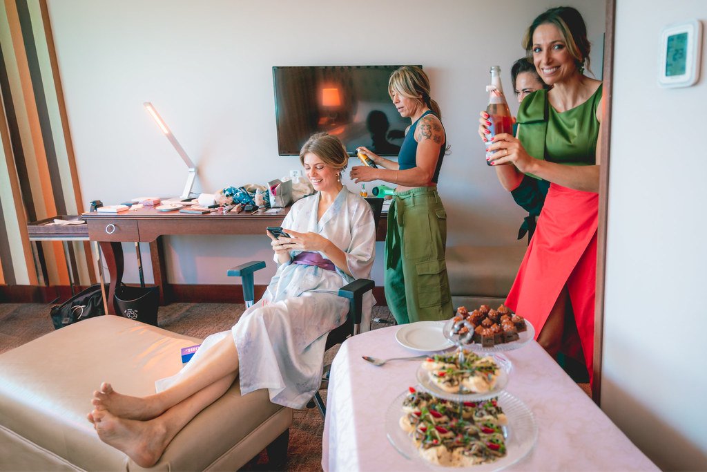 Mulheres se preparando em um quarto, uma sentada recebendo tratamento de beleza, outras de pé, uma segurando uma bebida com um sorriso, ao lado de uma mesa com petiscos.