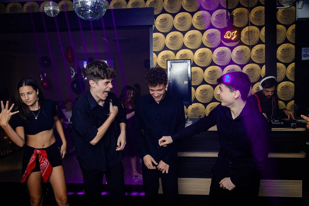 Grupo de jovens dançando e se divertindo em uma festa com decoração moderna e luzes coloridas, incluindo uma mulher com roupa de dança e três meninos sorrindo e dançando juntos.