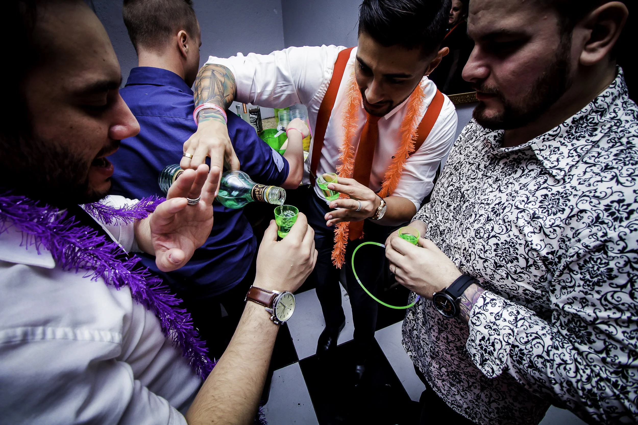Grupo de pessoas celebrando, vestidas com roupas festivas e acessórios coloridos, brindando com bebidas verdes em copinhos.