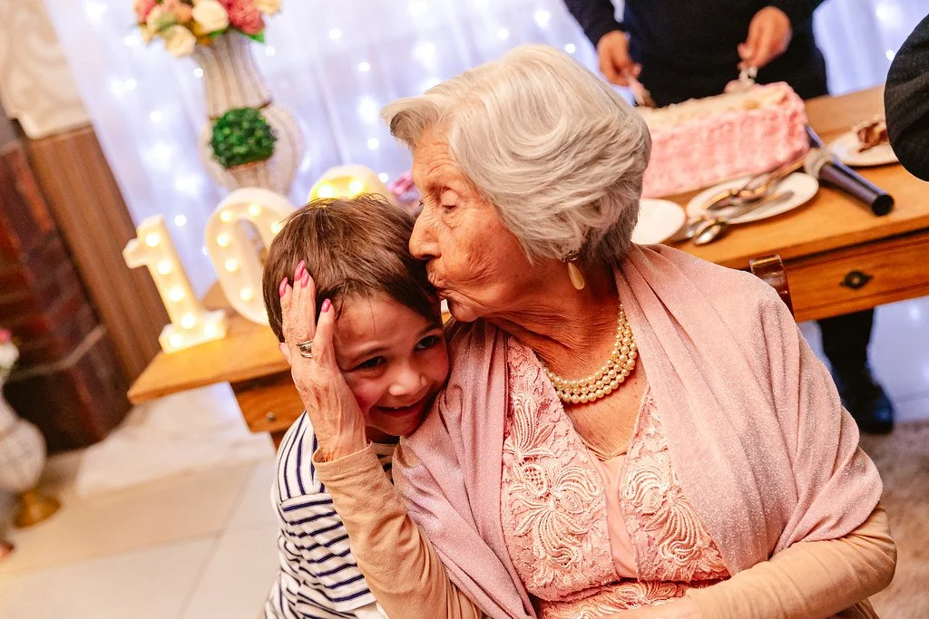 Uma avó idosa dá um beijo de agradecimento em um rosto de criança em uma festa de aniversário com decoração de luzes e um bolo ao fundo.