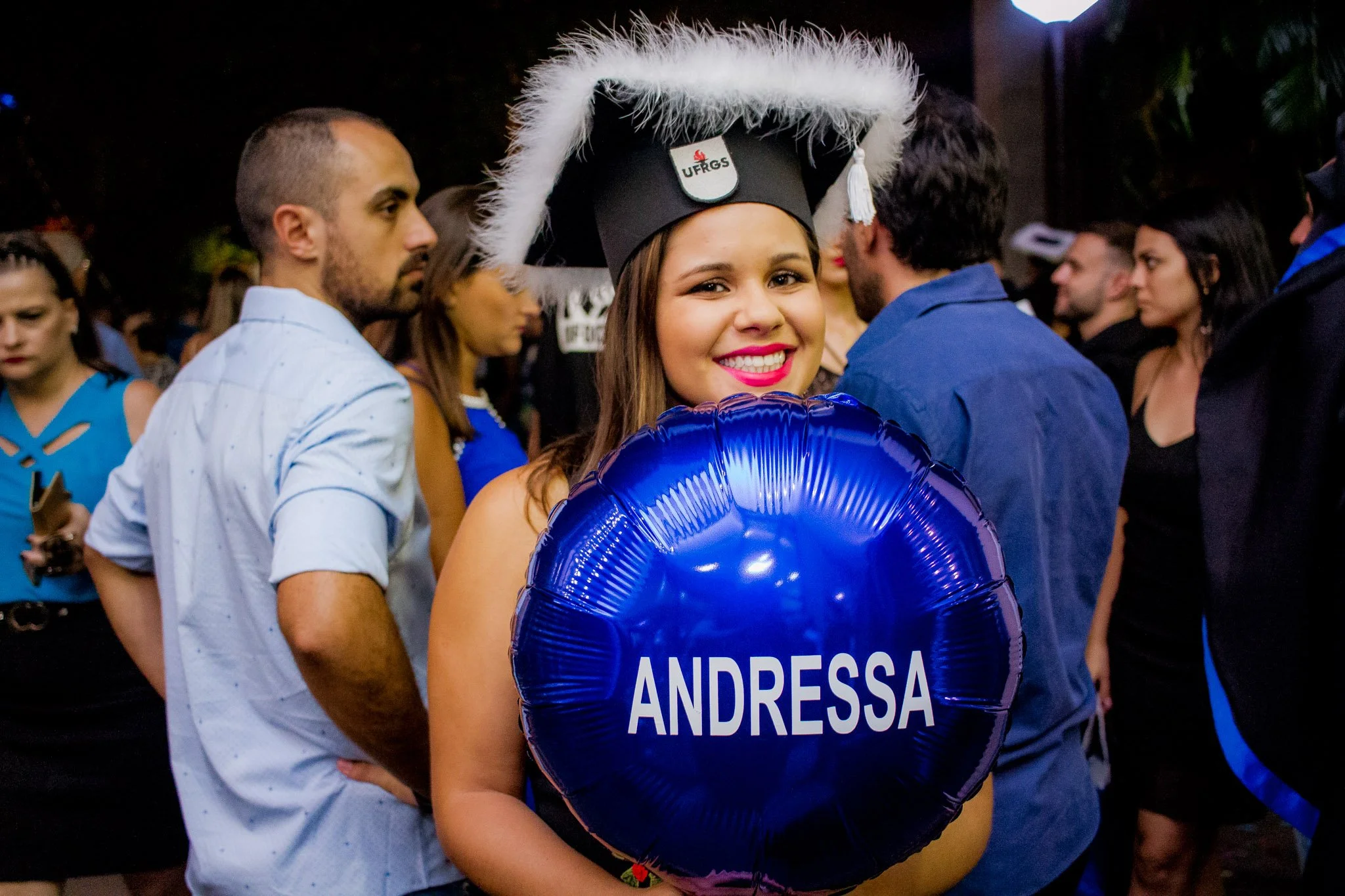 Jovem sorridente usando capela de formatura com a sigla UFRGS e segurando um balão oval azul com o nome 'Andressa', em uma cerimônia de formatura, rodeada de outros participantes.