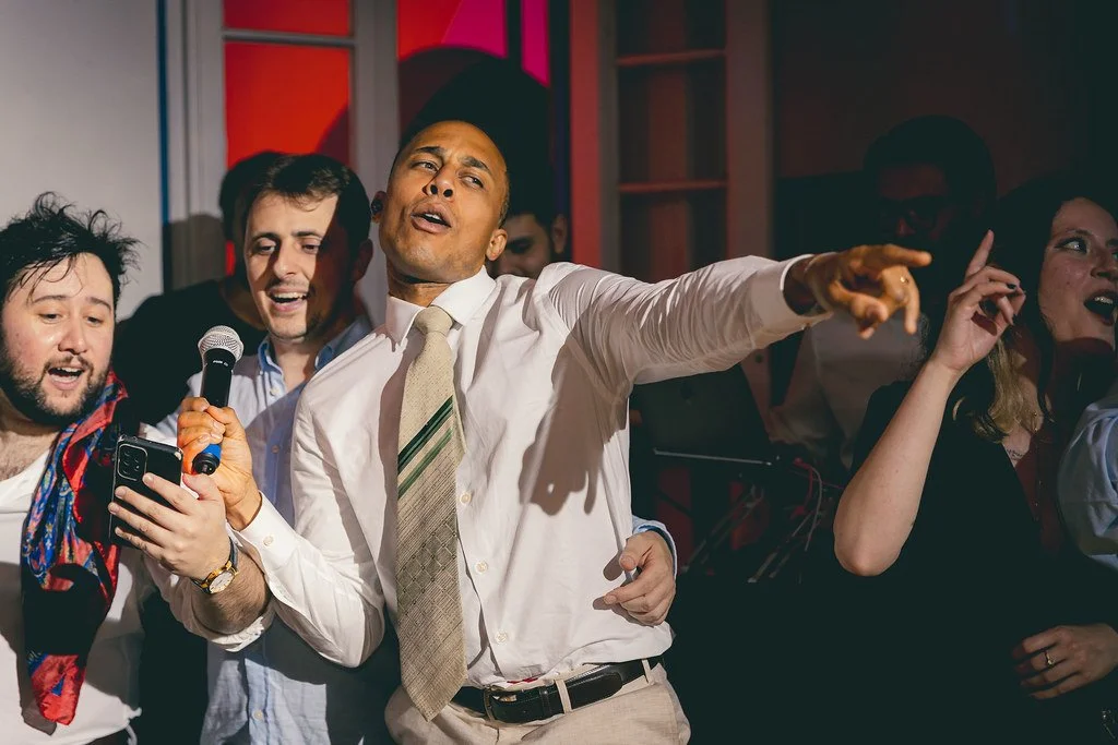 Homens e mulheres cantando e se divertindo em uma festa, com um homem de camisa branca, gravata e cabelo raspado apontando.