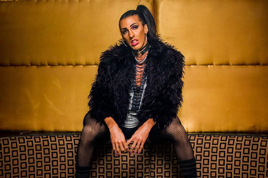 Mulher sentada em um sofá, vestindo um casaco de pelúcia preta, com cabelos presos em um rabo de cavalo, usando roupas negras e meias de rede, com uma parede dourada ao fundo.