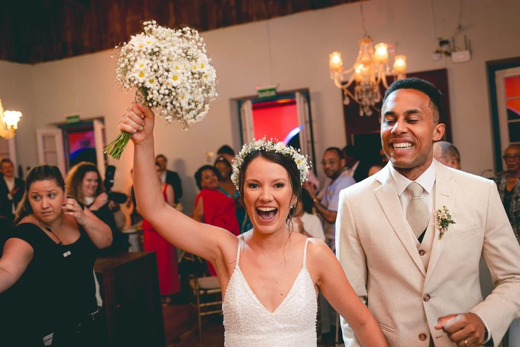Noiva sorridente segurando um buquê de flores brancas em uma festa de casamento, ao lado do noivo vestido com terno claro, rodeados por convidados em um ambiente decorado com lustres e janelas coloridas.
