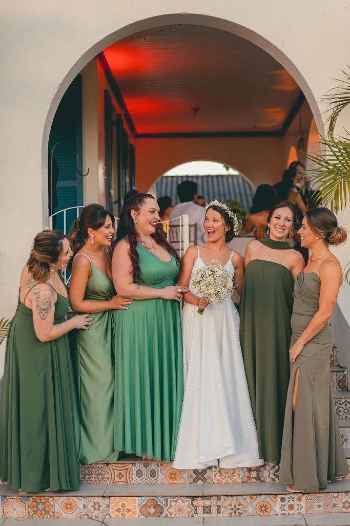 Grupo de mulheres em vestido de festa, com a noiva ao centro segurando um buquê, no estilo de uma cerimônia de casamento ao ar livre.