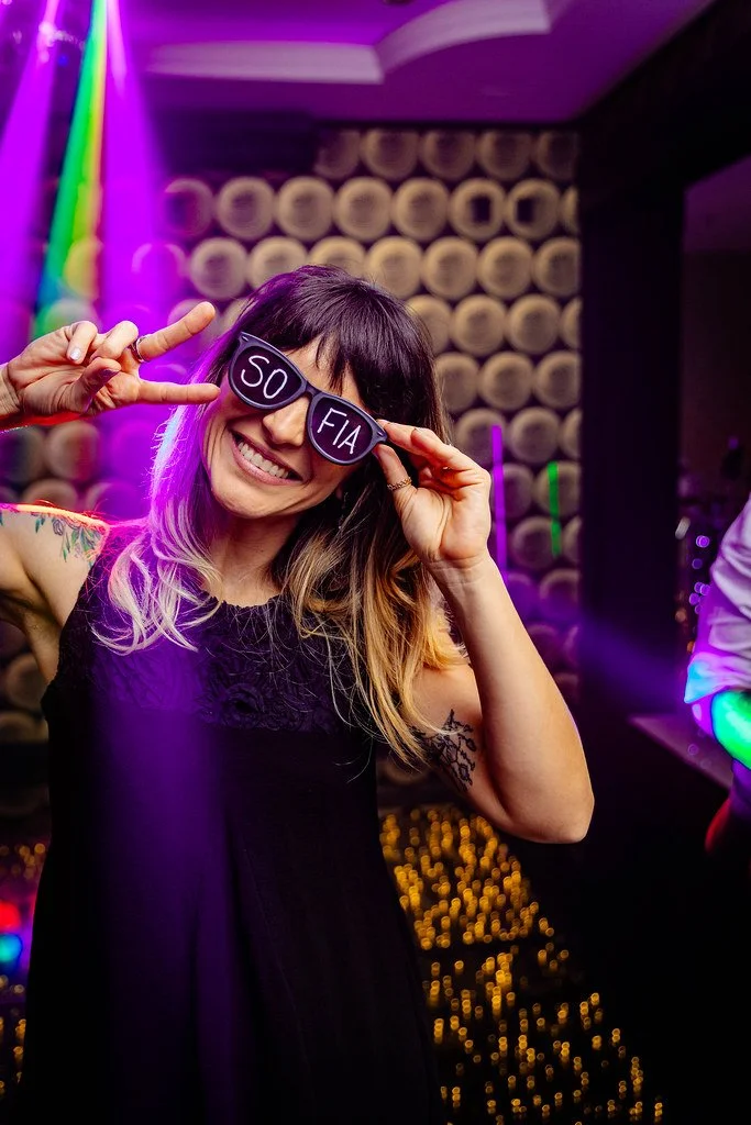 Mulher sorridente usando óculos de sol com a palavra ‘SO’ de um lado e ‘FIA’ de outro, fazendo o símbolo de paz com a mão, em ambiente com luzes coloridas e parede decorada