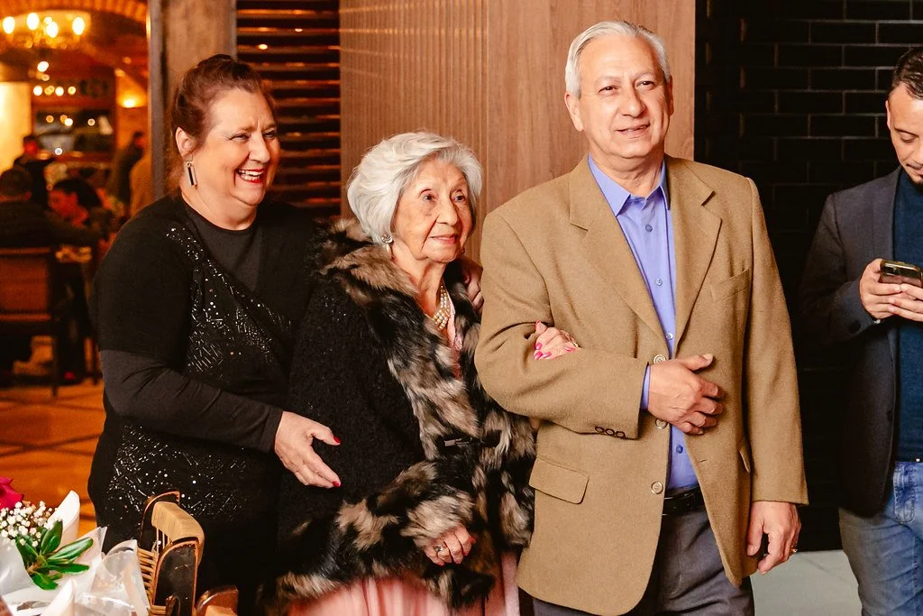 Quatro pessoas sorrindo e conversando em um ambiente interno, com duas mulheres idosas e um homem mais velho no centro, e um jovem à direita usando celular.