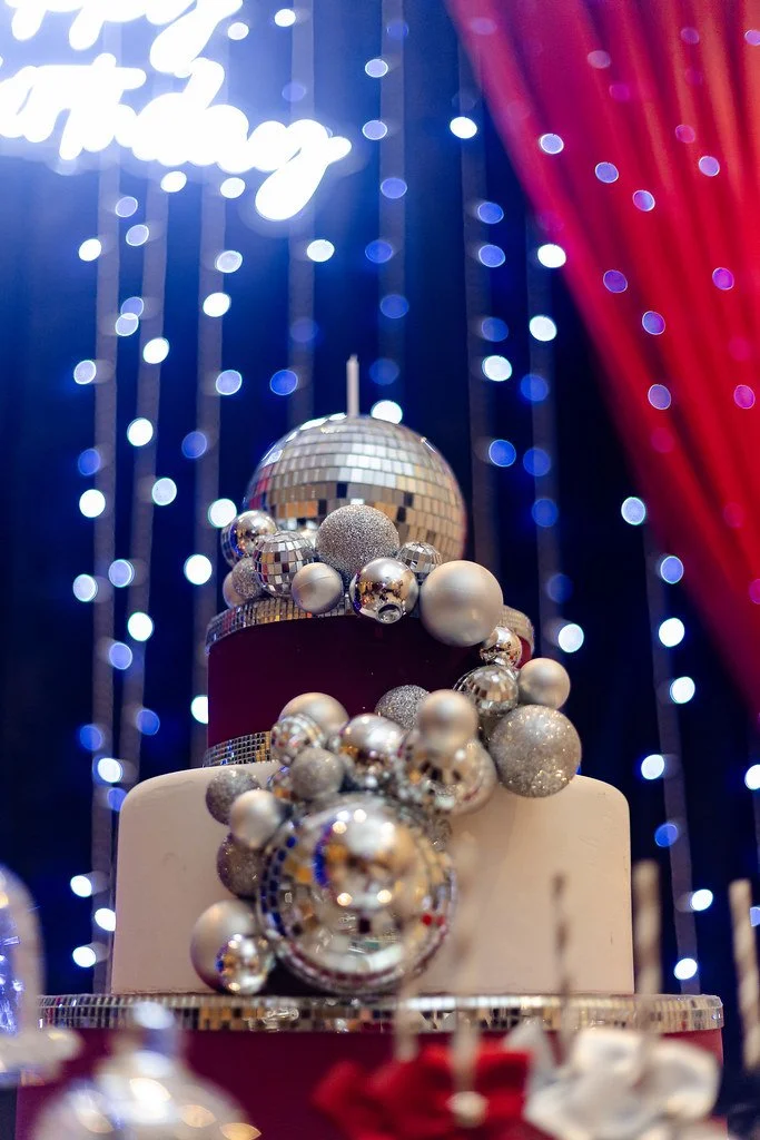 Bolo de festa decorado com bolas prateadas, douradas e prateadas em diferentes tamanhos, com fundo de luzes azuis e vermelho.