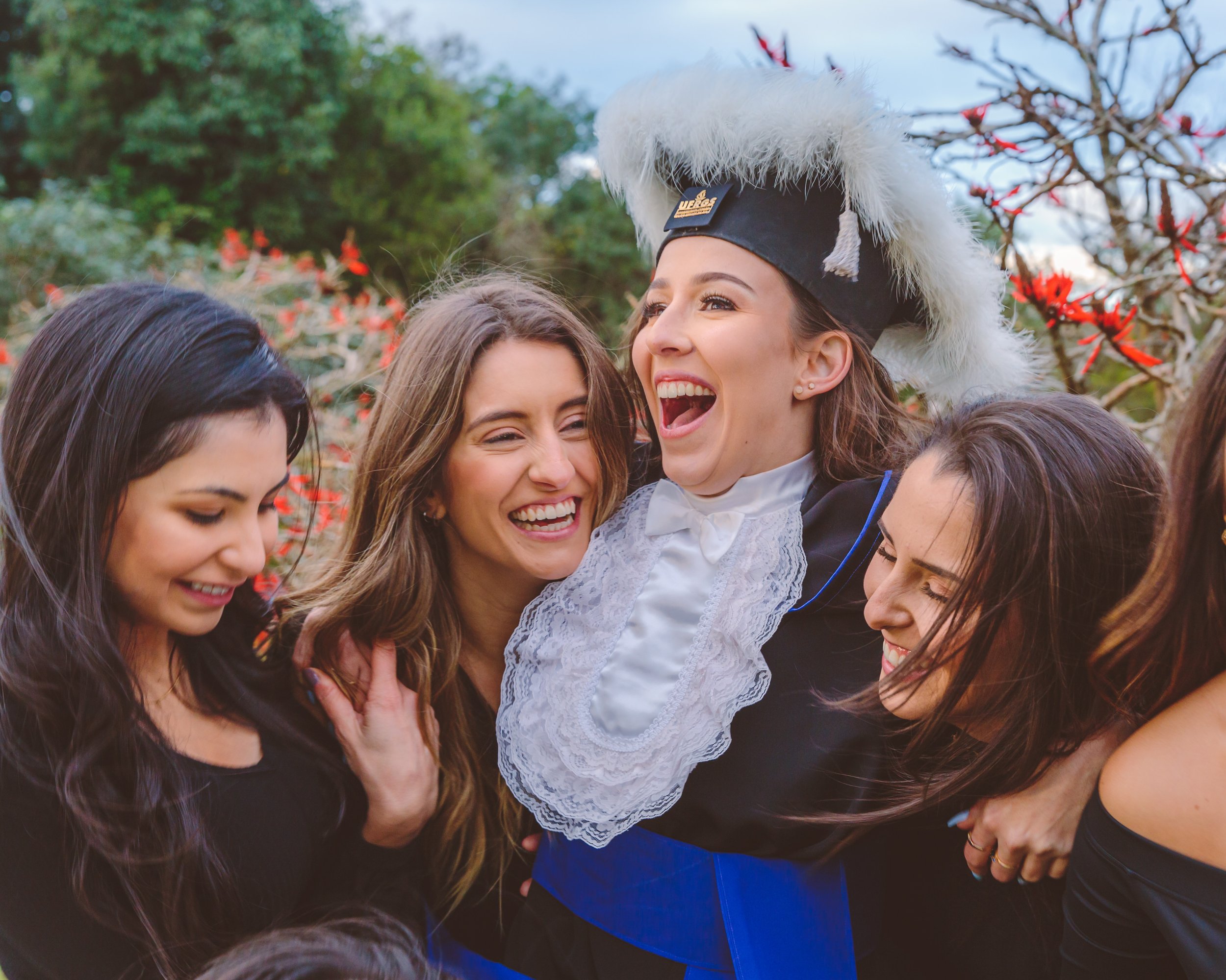 Mulher com chapéu de formatura sendo levantada por amigas em celebração ao ar livre com flores e árvores ao fundo.