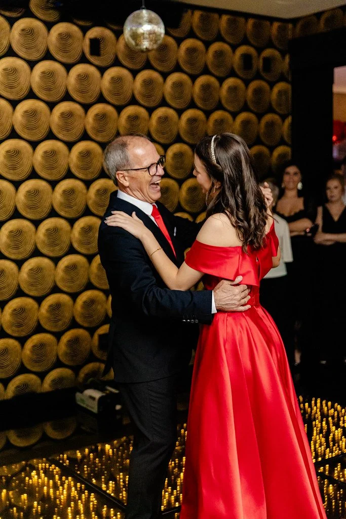 Homem e mulher dançando felizes em comemoração, com fundo decorado de cilindros de madeira e iluminação ambiente.
