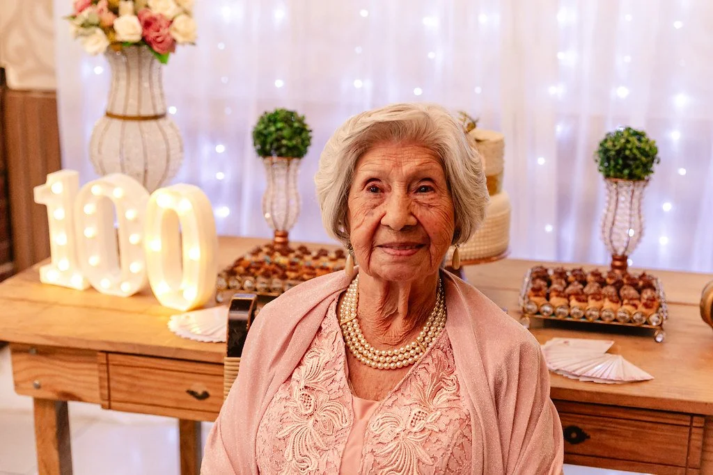 Uma senhora idosa sorrindo em uma festa de 100 anos, decorada com luzes brancas, vasos de flores, um bolo e doces na mesa ao fundo.