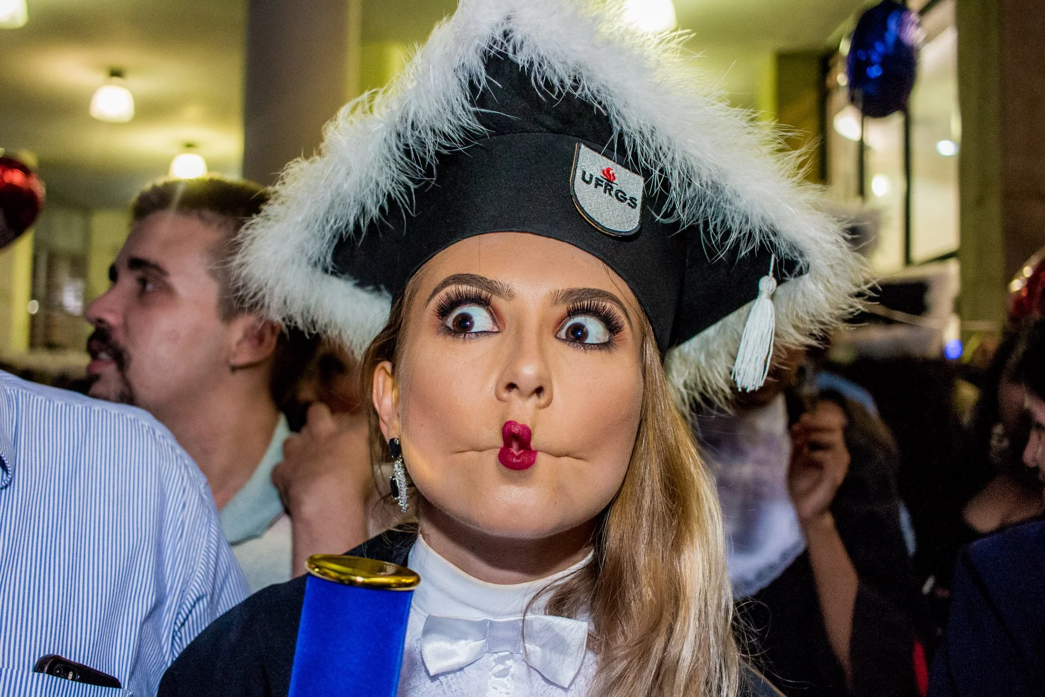Mulher com chapéu de formatura com plumas brancas, fazendo bico com expressão engraçada em uma festa com outras pessoas ao fundo.