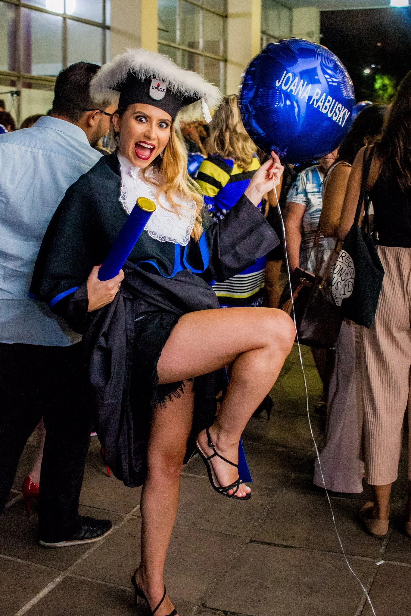 Mulher usando traje de formatura com capelo, segurando um diploma e um balão azul com o nome 'Joana Rabusky', em uma celebração de formatura com várias pessoas ao fundo.