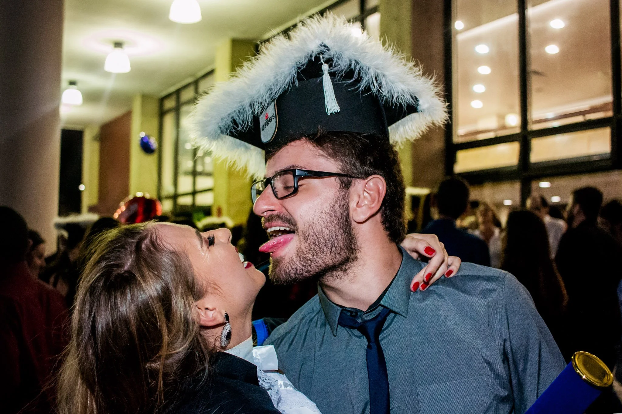 Homem com capelo de formatura e mulher em festa, ambos em posição próxima e expressão de diversão.