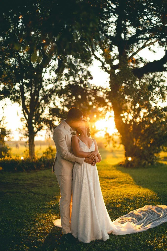 Casal de noivos em troca de olhares ao entardecer em um parque com árvores ao fundo.