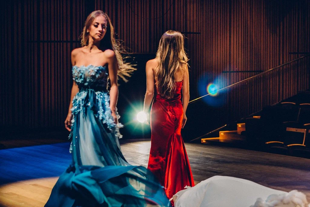 Duas mulheres em vestidos de noite elegantes em um palco de teatro escuro, uma com vestido azul e a outra com vestido vermelho, com um cenário de teatro ao fundo.