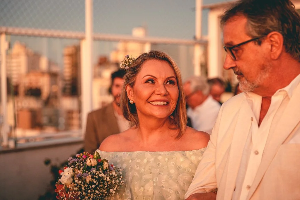Mulher sorrindo segurando um buquê de flores, em ambiente ao ar livre durante o pôr do sol, enquanto conversa com um homem de óculos.