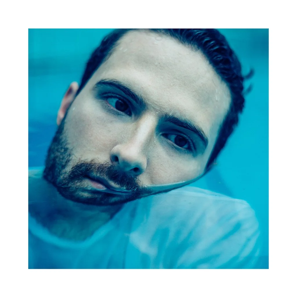 Retrato de um homem com barba e cabelos escuros, parcialmente submerso em água, com expressão séria, fundo azul.