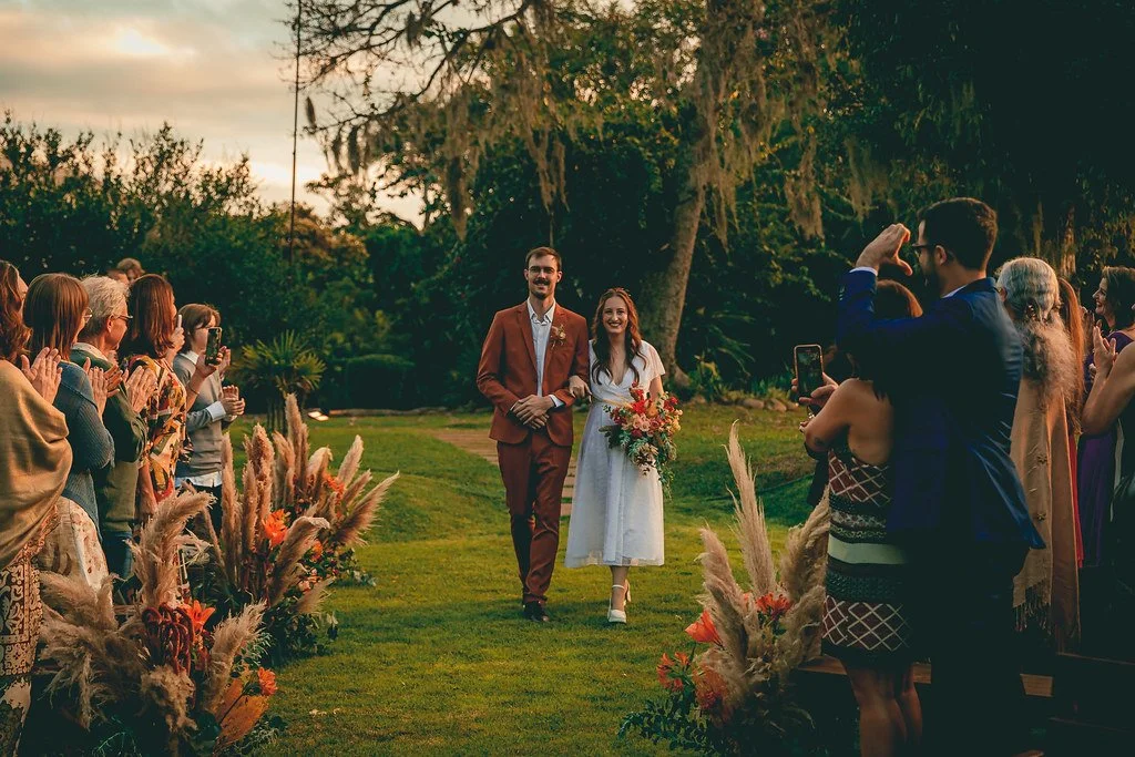 Casal de noivos caminhando pelo corredor no casamento ao ar livre, rodeados por amigos e familiares que celebram e tiram fotos, com decorações florais e vegetação natural ao fundo.