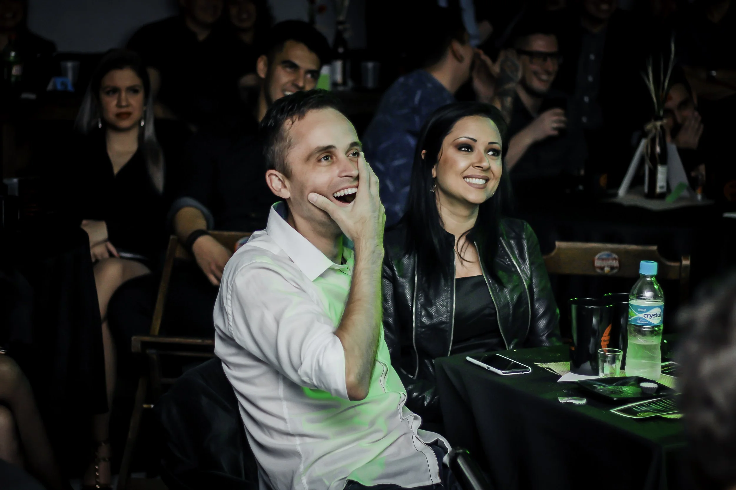 Pessoas assistindo a uma apresentação ou show, sorrindo e se divertindo, em ambiente com iluminação de palco.