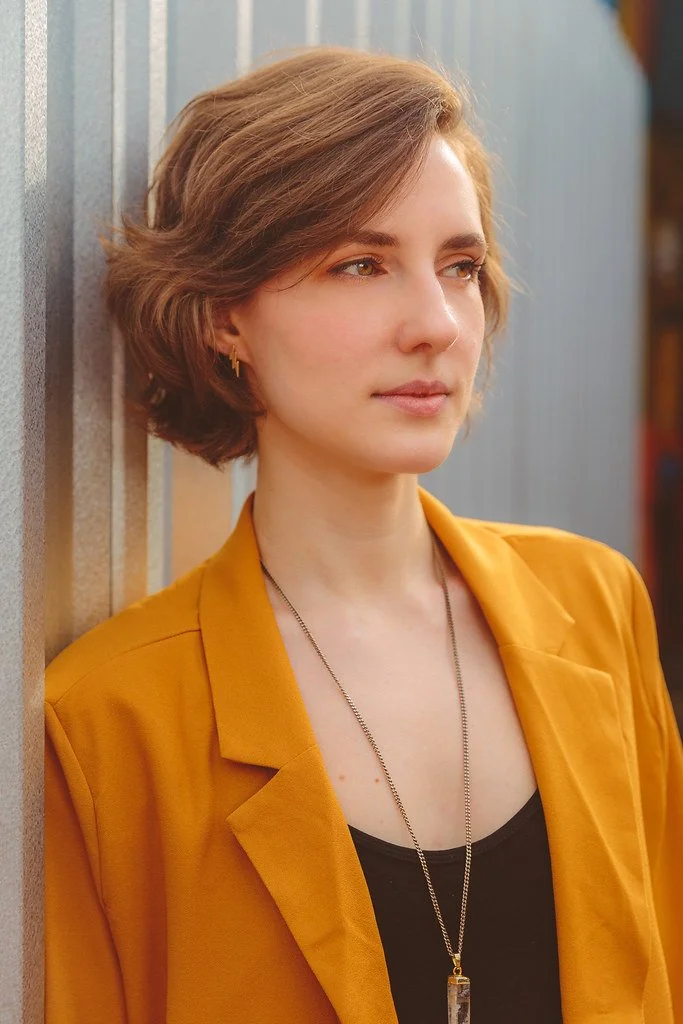 Mulher com cabelo castanho curto, vestindo blazer amarelo e camiseta preta, usando colar e brincos, apoiada contra uma parede de madeira.