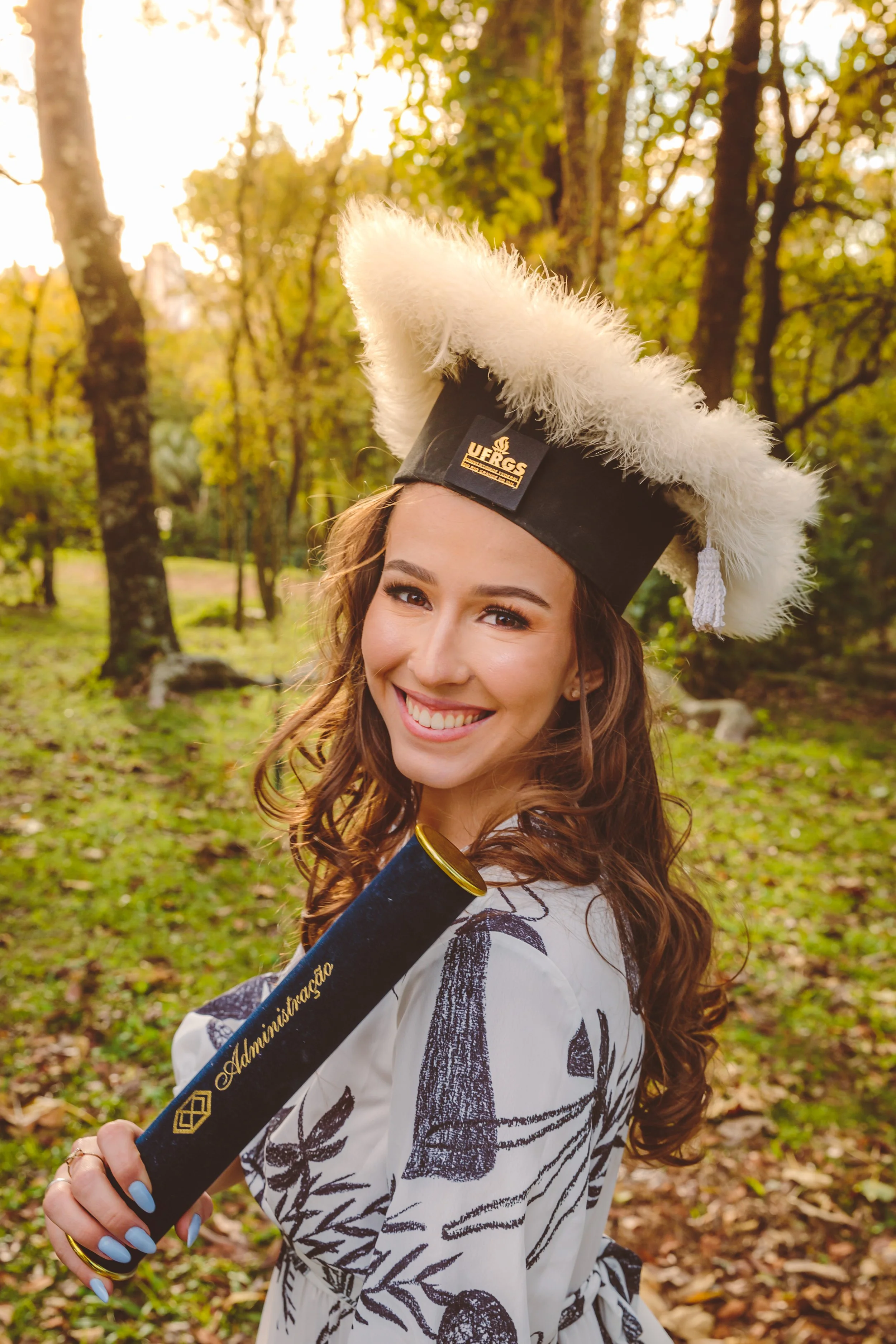 Jovem sorridente com cabelo castanho, usando chapéu de formatura com detalhes em branco, segurando um diploma de graduação, no parque durante o dia.