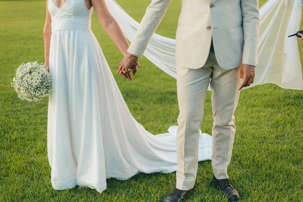Casal de casamento segurando as mãos, vestindo trajes formais em um campo verde, com uma cerimônia ao ar livre ao fundo.