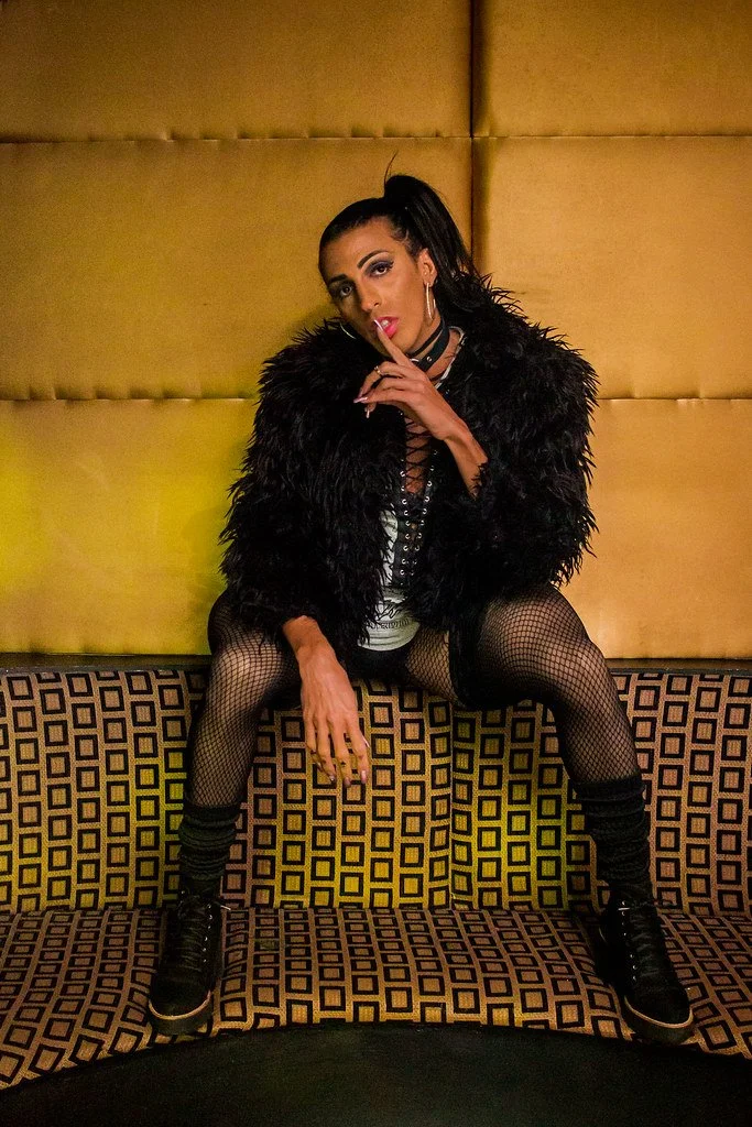 Mulher com cabelo preto em rabo de cavalo, vestindo jaqueta de pelos pretos, meias de rede, vestido branco e tênis pretos, sentada em um banco com fundo dourado.