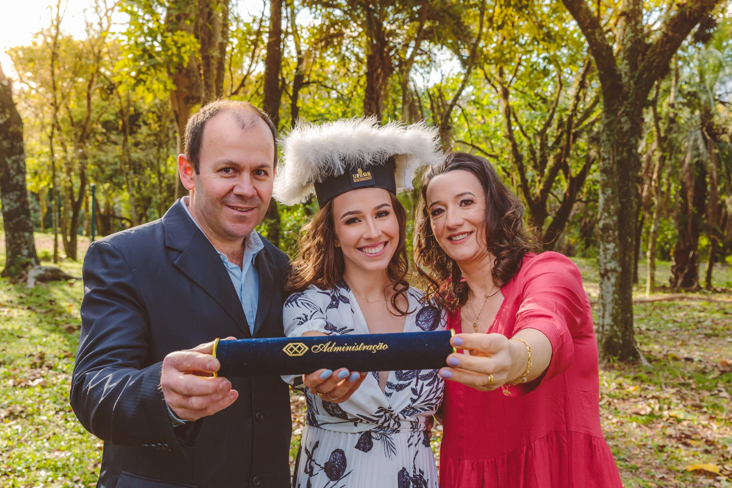 Três pessoas sorrindo em uma floresta, uma delas usando capelo de formatura, segurando um diploma, ao lado de uma mulher em um vestido vermelho e um homem de terno, celebrando uma conquista acadêmica ou profissional.